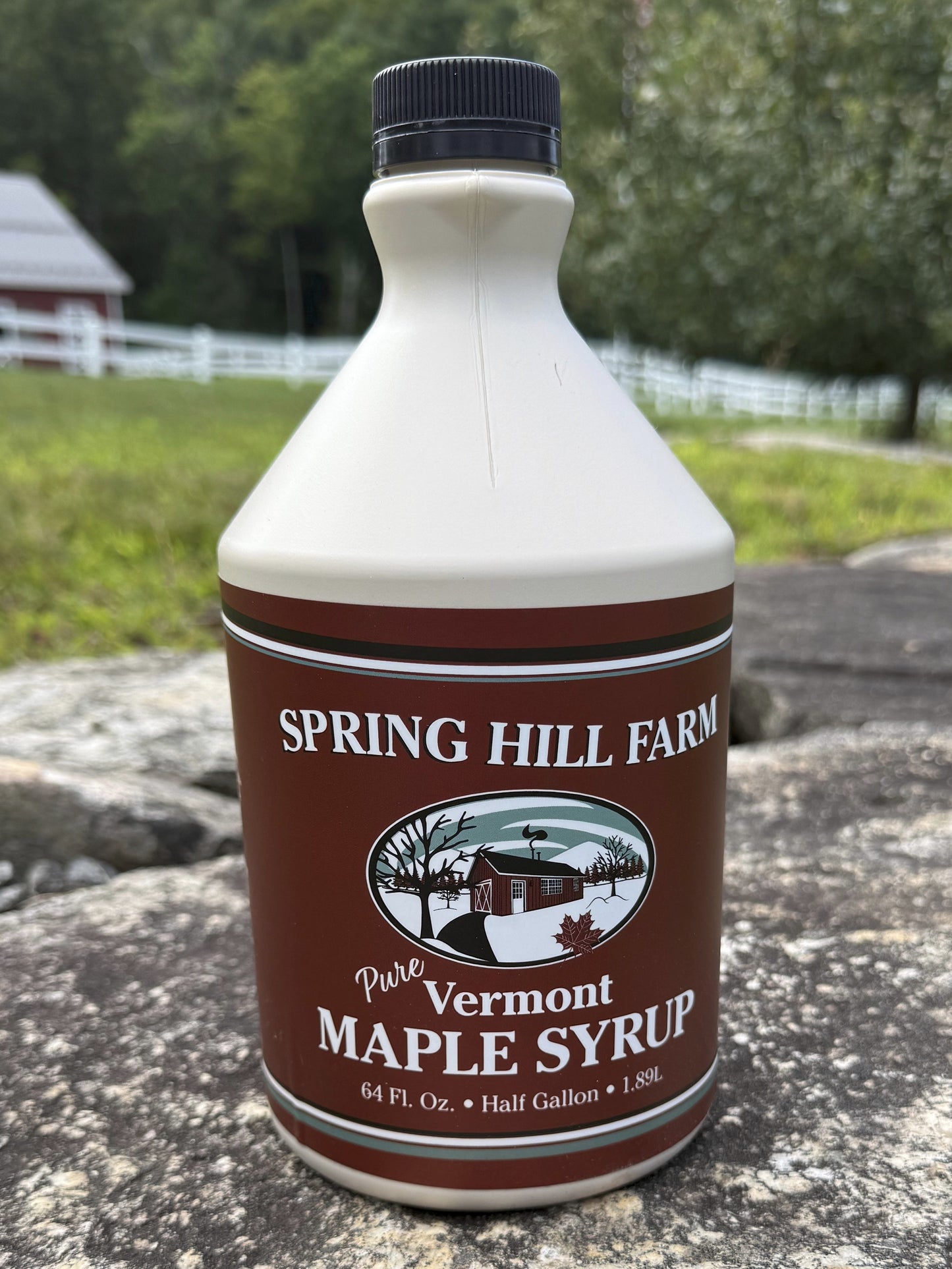 A half-gallon bottle of Spring Hill Farm Vermont Maple Syrup placed on a large stone outdoors, with a grassy field, trees, and a barn in the background.