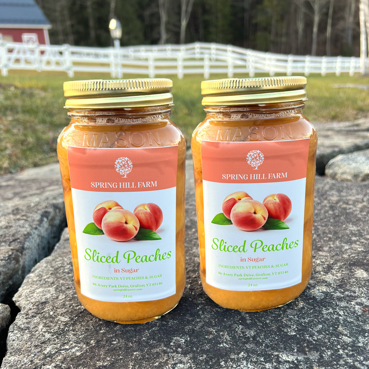two mason jars of spring hill farm sliced peaches in sugar sitting on a stone wall in front of a field and white fence