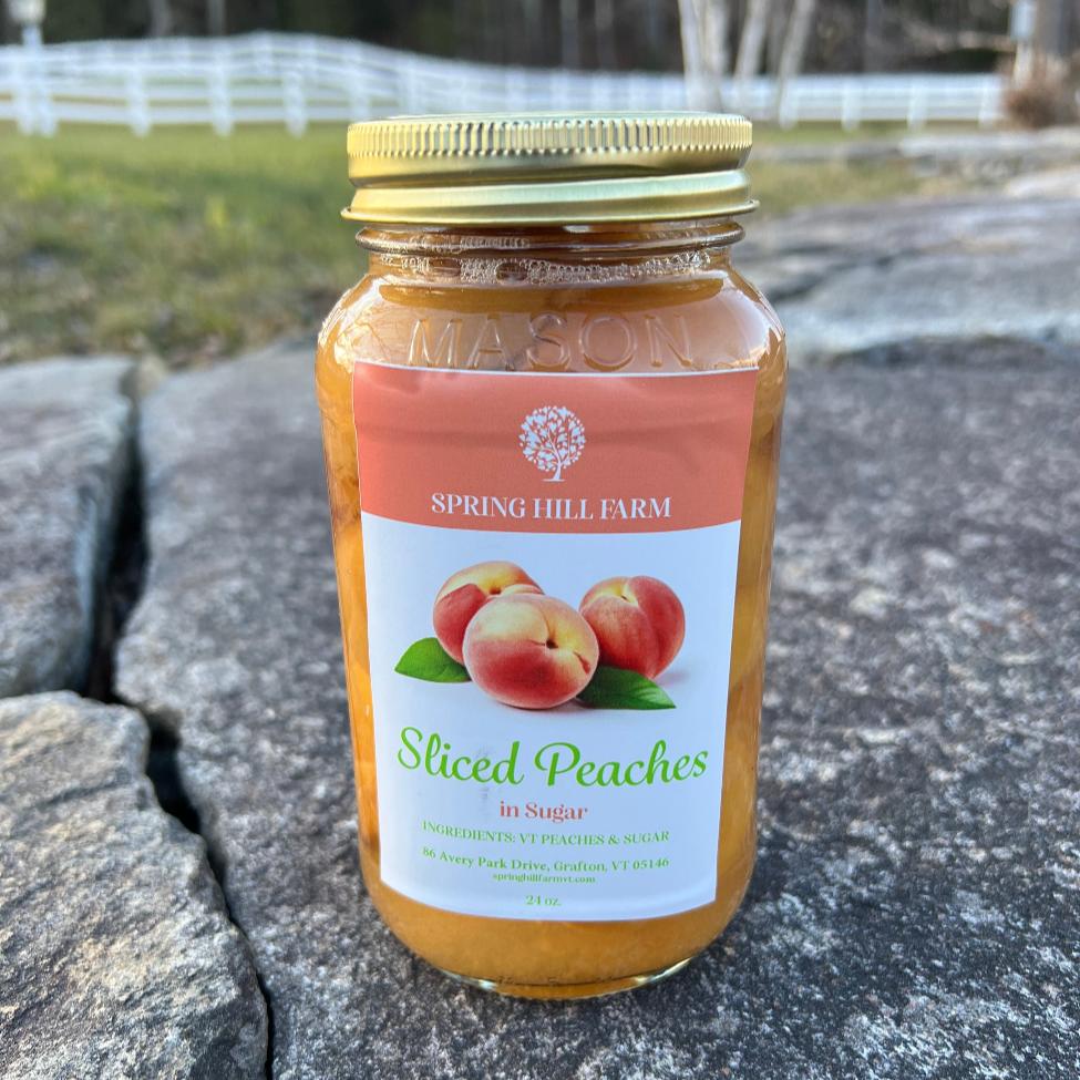 mason jar of spring hill farm sliced peaches in sugar sitting on a stone wall in front of a field and white fence